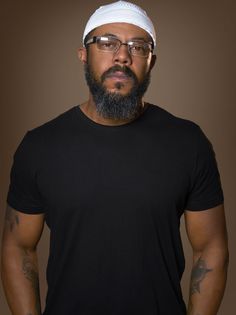 a man with glasses and a beard wearing a black t - shirt stands in front of a brown background