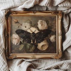 a butterfly sitting on top of a white blanket next to some flowers and leaves in a gold frame