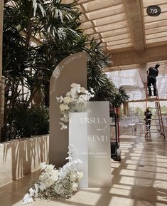 a tall white sign sitting in the middle of a room next to a plant filled with flowers