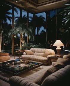 a living room filled with furniture and large windows at night, next to palm trees