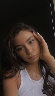 a woman in white tank top holding her hand to her head and looking at the camera