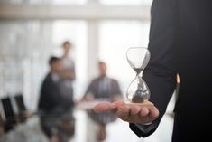 a man holding an hourglass in his hand while other people are sitting at desks behind him