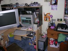 a cluttered desk with a computer monitor and keyboard on it in front of a bookshelf