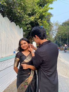 a man fixing the ear of a woman in a black sari, standing next to a white fence