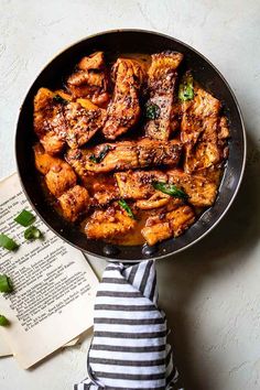 a skillet filled with chicken wings and garnished with green onions next to an open book