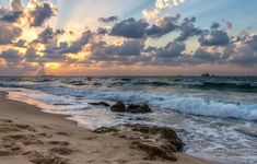 the sun is setting over the ocean with waves crashing on the beach and rocks in the foreground