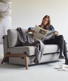 a woman sitting on a gray couch reading a newspaper while holding a coffee cup and looking at the camera