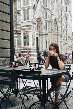 a woman sitting at an outdoor table drinking from a cup in front of a building