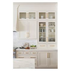 a kitchen with white cabinets and marble counter tops, gold pulls on the glass doors