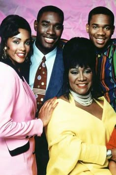 a group of people standing next to each other in front of a pink background with one woman wearing a yellow dress