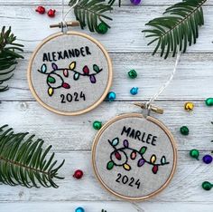 two embroidered christmas ornaments hanging on a white wooden table with pine needles and colorful beads