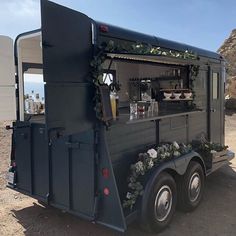 the back end of a black food truck parked on top of a dirt field next to a mountain