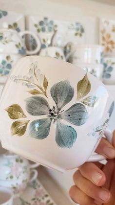 a hand holding a white and green flowered cup in front of plates on the wall