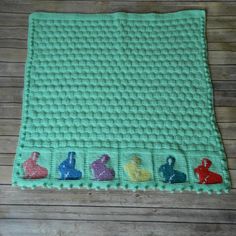 a green crocheted blanket with small rabbits on the front and sides, sitting on a wooden floor