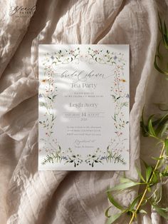 the wedding card is laying on top of a bed with white sheets and greenery