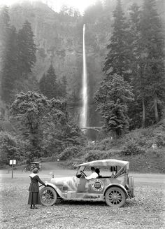 an old car parked in front of a waterfall with a woman standing next to it