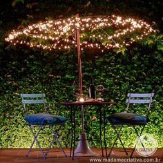 two chairs sitting under an umbrella with lights on it in front of a hedge wall