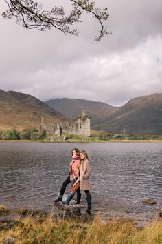 two people standing on the shore of a lake in front of a castle and mountains