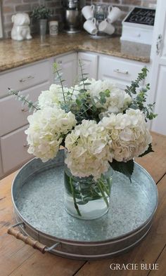 white flowers are in a glass vase on a metal platter at the kitchen table