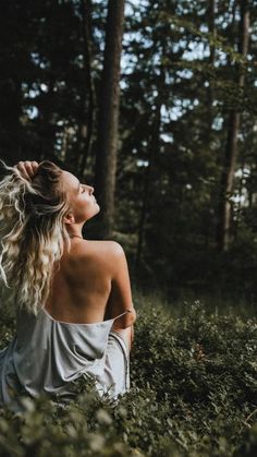 a woman sitting in the middle of a forest looking up into the sky with her hair blowing back