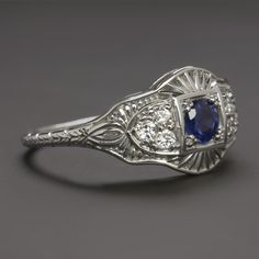 This beautiful and unique ring features a richl blue sapphire set in a diamond studded vintage style design that will never go out of style! The sapphire has a richly saturated blue hue that contrasts strikingly with the 14k white gold setting. Utterly classic and romantic, the antique style setting is richly textured with intricate filigree and milgrain. The effect is chic but also dreamy and softly feminine, a perfect choice for a timelessly stylish right hand ring or alternative engagement ri Diamond And Blue Sapphire Wedding Ring, Classic Blue Diamond Ring, Classic Blue Diamond Ring With Accents, Blue Single Cut Diamond Jewelry In Platinum, Platinum Jewelry With Blue Single Cut Diamonds, Blue Platinum Jewelry With Single Cut Diamonds, Vintage Platinum Diamond Ring With Gemstone, Classic Formal Diamond Ring With Lab-created Sapphire, Art Deco Blue Diamond Ring With Center Stone