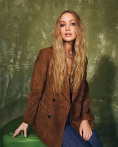 a woman sitting on top of a green suitcase wearing a brown jacket and blue jeans