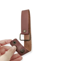 a hand holding a brown leather belt on top of a white surface with a metal buckle