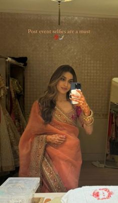 a woman in an orange sari taking a selfie with her cell phone while looking into the mirror