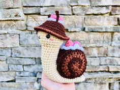 a small crocheted snail with a hat on it's head is held in front of a brick wall