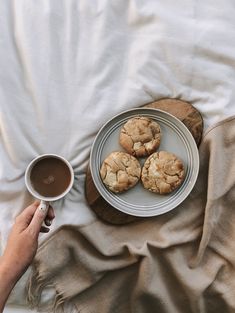 a person holding a cup of coffee next to some cookies on a plate and blanket