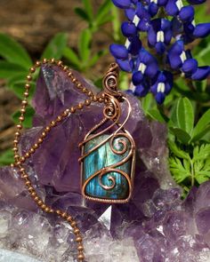 a wire wrapped pendant sitting on top of purple rocks next to blue flowers and greenery