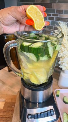 a person holding a lemon wedge over a blender filled with cucumber and lemon slices