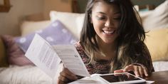 a woman sitting on a bed reading a book and smiling at the camera while holding papers in her hands