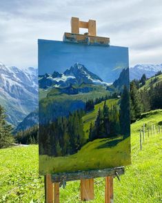 an easel with a painting on it sitting in the grass near mountains and trees