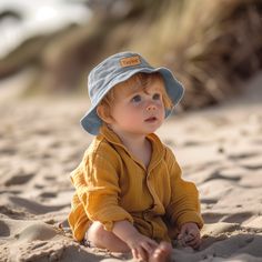 a little boy sitting in the sand wearing a hat