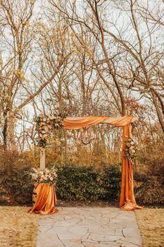 an outdoor ceremony with orange draping and flowers on the arch, surrounded by trees