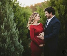 a pregnant couple standing next to each other in the middle of some trees at sunset