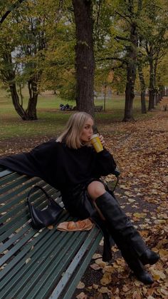 a woman sitting on a park bench with her legs crossed and drinking from a cup