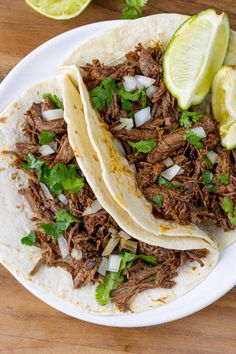 two shredded beef tacos on a white plate with limes and cilantro