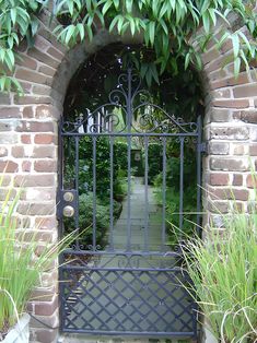 an iron gate in the middle of a brick wall with grass growing around it and bushes on either side