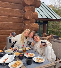 three women taking a selfie at a table with food and drinks in front of a log cabin