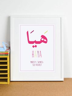 a white framed print with the words hayya in pink and yellow, on a shelf next to a stack of books