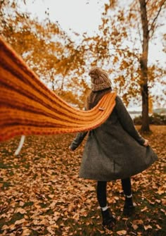 a woman is walking through the leaves with her long orange scarf in front of her