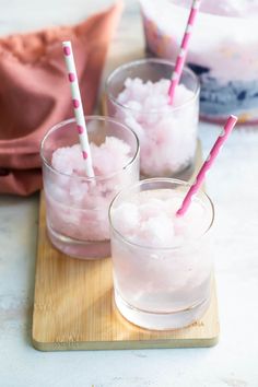 three glasses filled with ice sitting on top of a wooden cutting board next to two pink straws