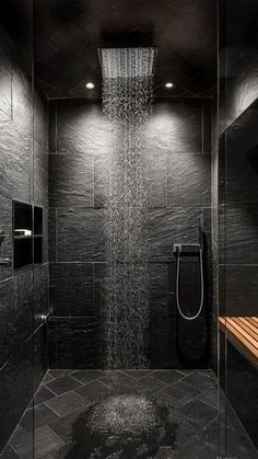 a walk in shower sitting next to a wooden bench and tiled walls with black tiles