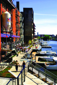 people are walking on the boardwalk near boats docked in the water and red brick buildings