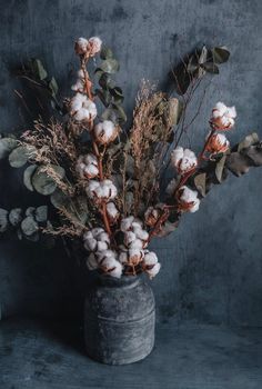 a vase filled with cotton and greenery on top of a wooden table next to a gray wall