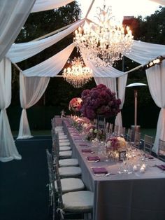 a long table is set up with white drapes and chandeliers for an outdoor event