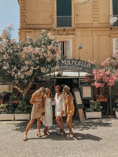 three women standing in front of a building with pink flowers on the outside and green shutters