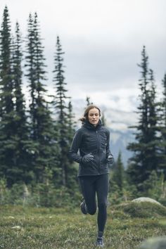 a woman running in the woods on a cloudy day with mountains and trees behind her